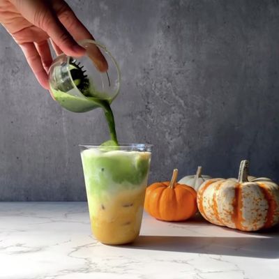 Matcha being poured into a pumpkin-spice milk drink with fall pumpkins